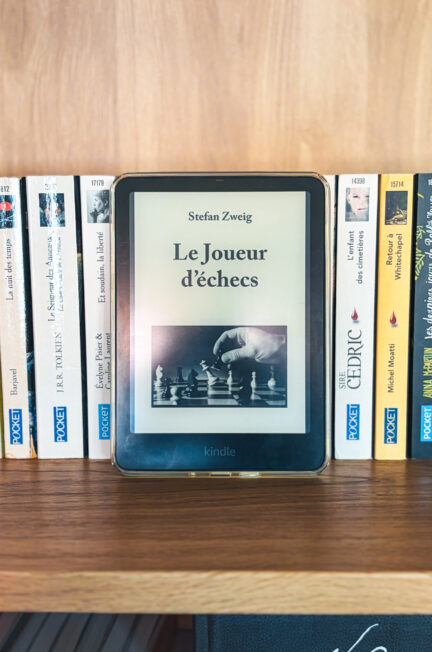 Liseuse Kindle affichant la couverture du livre Le joueur d'échecs posée sur une bibliothèque en bois clair devant des livres de poches de la collection Pocket.