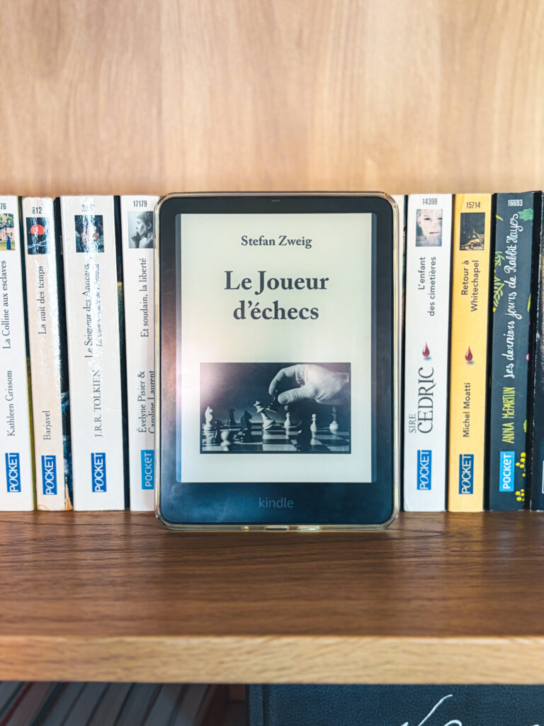 Liseuse Kindle affichant la couverture du livre Le joueur d'échecs posée sur une bibliothèque en bois clair devant des livres de poches de la collection Pocket.
