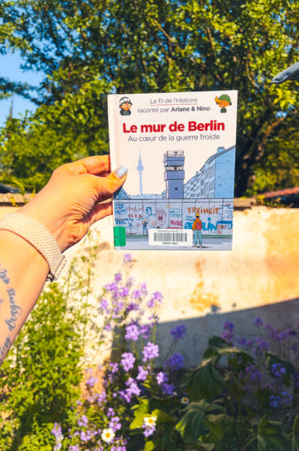 Le livre Le mur de Berlin au coeur de la guerre froide de Fabrice Erre et Sylvain Savoia tenu à la main dans un jardin fleuri au printemps.