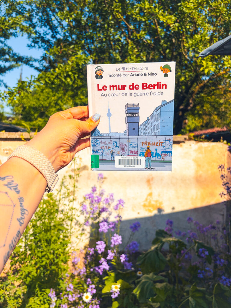 Le livre Le mur de Berlin au coeur de la guerre froide de Fabrice Erre et Sylvain Savoia tenu à la main dans un jardin fleuri au printemps.