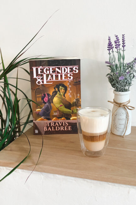 Le roman Légendes et Lattes de Travis Baldree posé sur une étagère en bois à côté d’un latte macchiato mousseux et d’un vase de lavande, dans une ambiance cosy et chaleureuse.