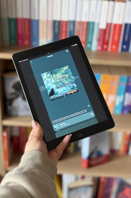 Main tenant une tablette affichant un lecteur audio avec Leibhaftig de Christa Wolf, devant une bibliothèque remplie de livres aux tranches colorées.
