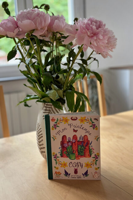 Le livre Mon printemps cosy de Caroline Millet posé sur une table en bois devant un vase rempli de pivoines roses, dans une ambiance printanière lumineuse et apaisante.