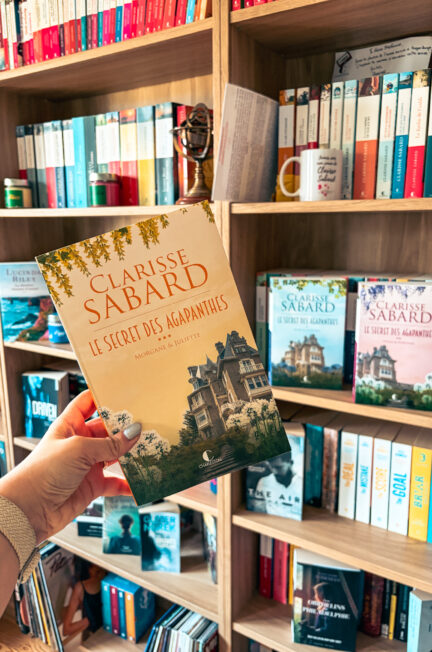Le roman Morgane et Juliette de Clarisse Sabard tenu devant une grande bibliothèque remplie de livres, avec plusieurs tomes de la saga Le secret des Agapanthes visibles en arrière-plan