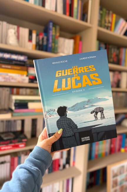 Episode II Les guerres de Lucas de Renaud Roche et Laurent Hopman tenu à la main devant une bibliothèque remplie de livres, avec la couverture représentant George Lucas face à un paysage enneigé inspiré de l'Empire contre-attaque.