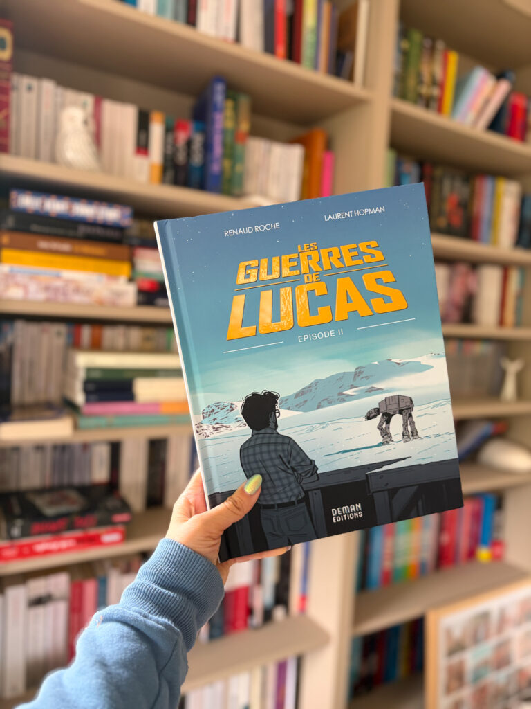 Episode II Les guerres de Lucas de Renaud Roche et Laurent Hopman tenu à la main devant une bibliothèque remplie de livres, avec la couverture représentant George Lucas face à un paysage enneigé inspiré de l'Empire contre-attaque.