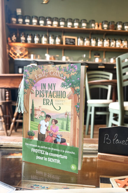 In my pistachio Era - Juliette’s Version d’Emily Collins posé sur une table dans le salon de thé Fleurs de Saison à Douarnenez, devant un mur garni d’étagères remplies de bocaux.