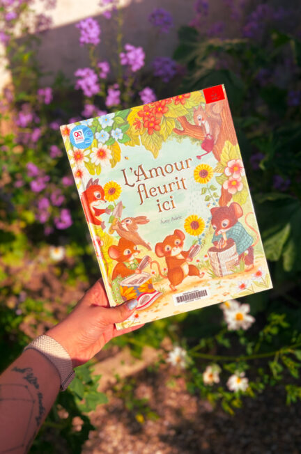 Le livre L’amour fleurit ici d’Amy Adele présenté dans un jardin fleuri au printemps, entouré de fleurs violettes et de marguerites.