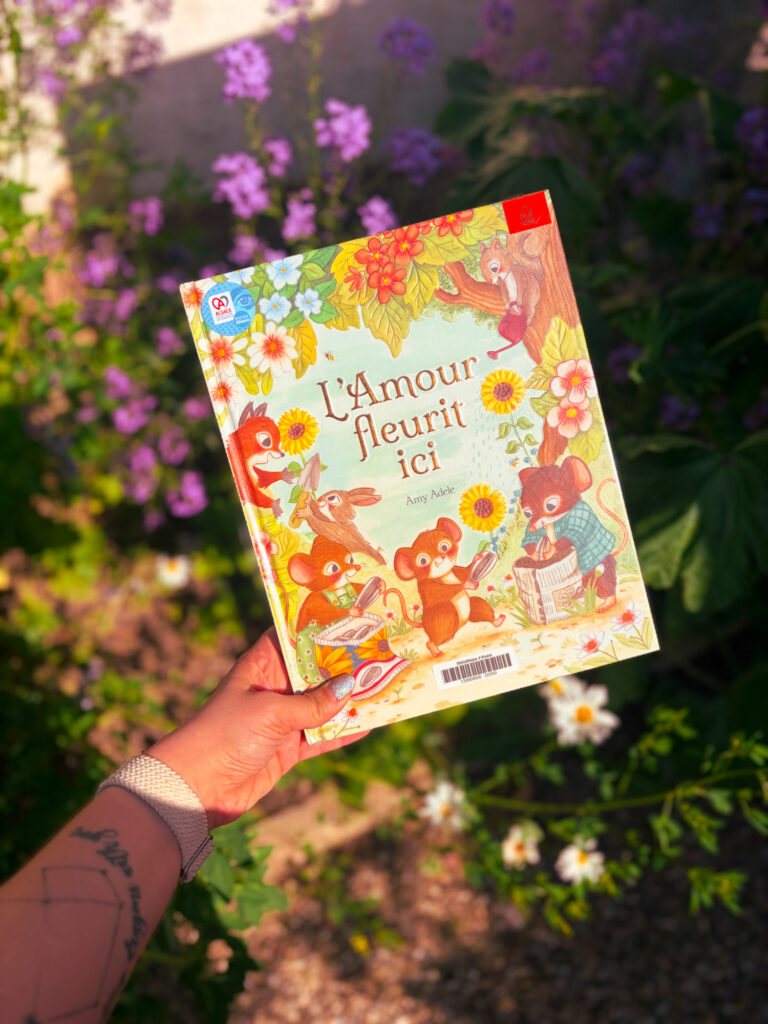 Le livre L’amour fleurit ici d’Amy Adele présenté dans un jardin fleuri au printemps, entouré de fleurs violettes et de marguerites.