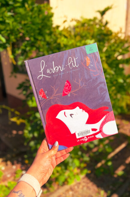 Le livre L’arbre-lit de Silène Edgar et Gilles Freluche est tenu à la main dans un jardin baigné de soleil, avec une végétation verte floue en arrière-plan.