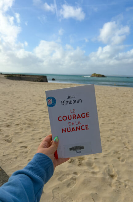 Le courage de la nuance de Jean Birnbaum tenu à la main sur la plage de Tréboul à Douarnenez, face à la mer sous un ciel bleu parsemé de nuages. 