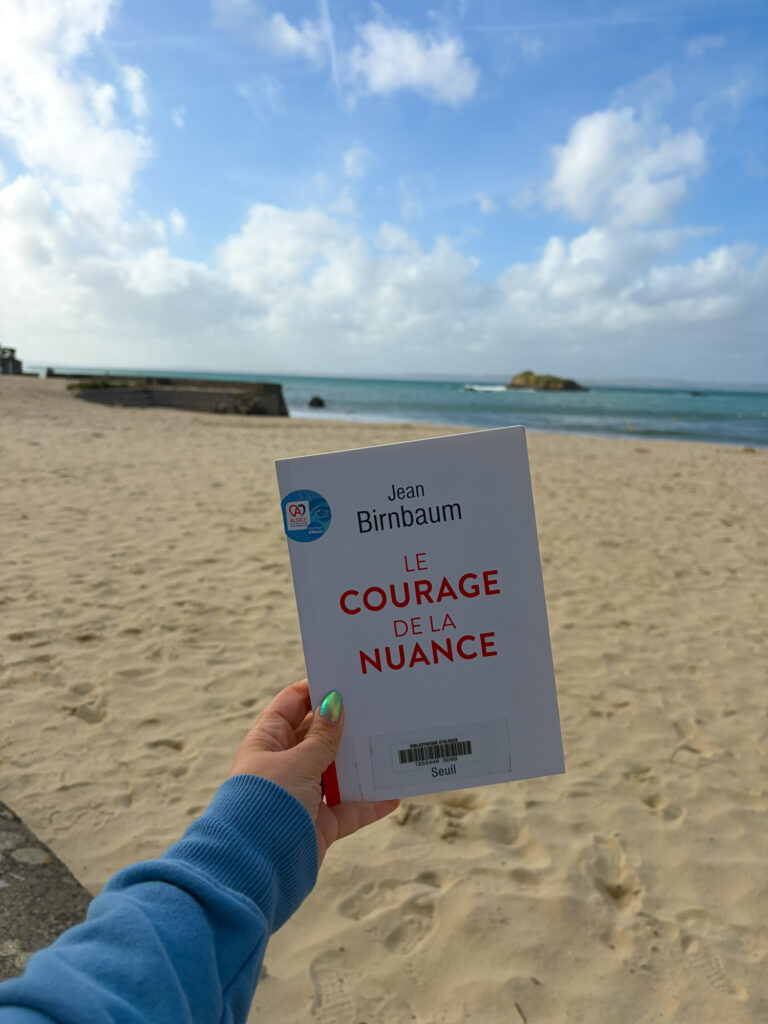 Le courage de la nuance de Jean Birnbaum tenu à la main sur la plage de Tréboul à Douarnenez, face à la mer sous un ciel bleu parsemé de nuages. 