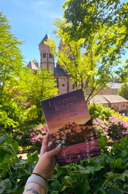 Le roman Le domaine de l’héritière de Lucinda Riley est tenu à la main devant les jardins fleuris et l’abbaye de Maria Laach, sous un ciel bleu éclatant.