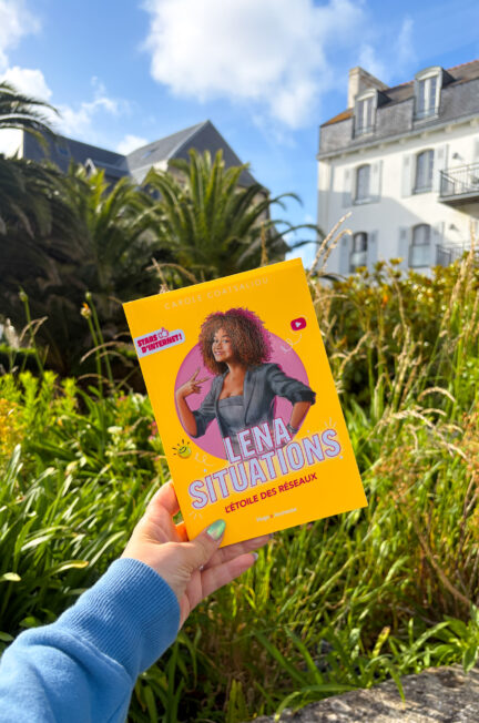 Le livre L’étoile des réseaux de Carole Coatsaliou tenu à la main devant un jardin luxuriant avec des palmiers et une maison blanche sous un ciel bleu. Il s'agit de l'hôtel Valdys Resort de Douarnenez.