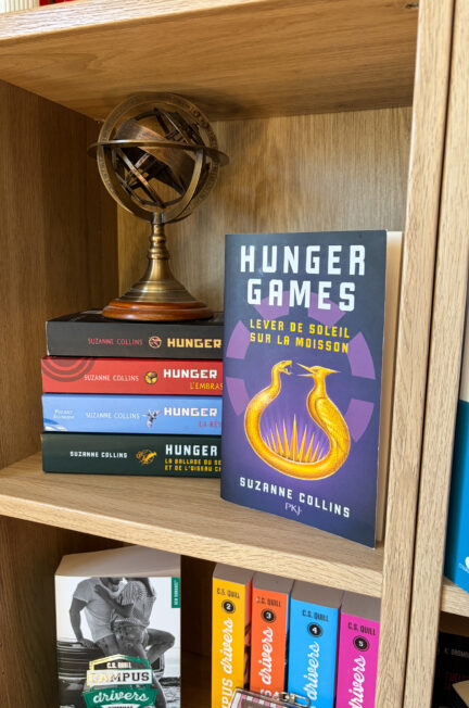 Le roman Lever de soleil sur la moisson de Suzanne Collins est posé sur une étagère en bois, devant les autres tomes de la saga Hunger Games et une sphère armillaire décorative.
