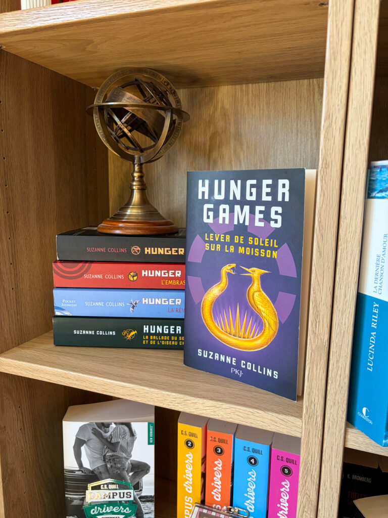 Le roman Lever de soleil sur la moisson de Suzanne Collins est posé sur une étagère en bois, devant les autres tomes de la saga Hunger Games et une sphère armillaire décorative.
