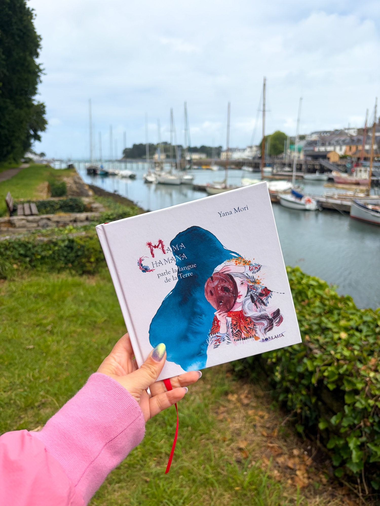 Le livre Mama Chamana parle la langue de la Terre de Yana Mori tenu à la main devant le port de Douarnenez en Bretagne, avec les voiliers amarrés en arrière-plan sous un ciel nuageux.