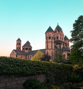 Abbaye : Vue extérieure de l’abbaye bénédictine de Maria Laach au coucher du soleil derrière un mur couvert de végétation.