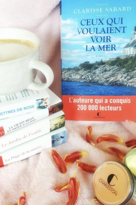 Le roman Ceux qui voulaient voir la mer de Clarisse Sabard est photographié à côté d'une tasse de café, de plusieurs romans de l'autrice, de bougies Charleston et de pétales de fleurs.