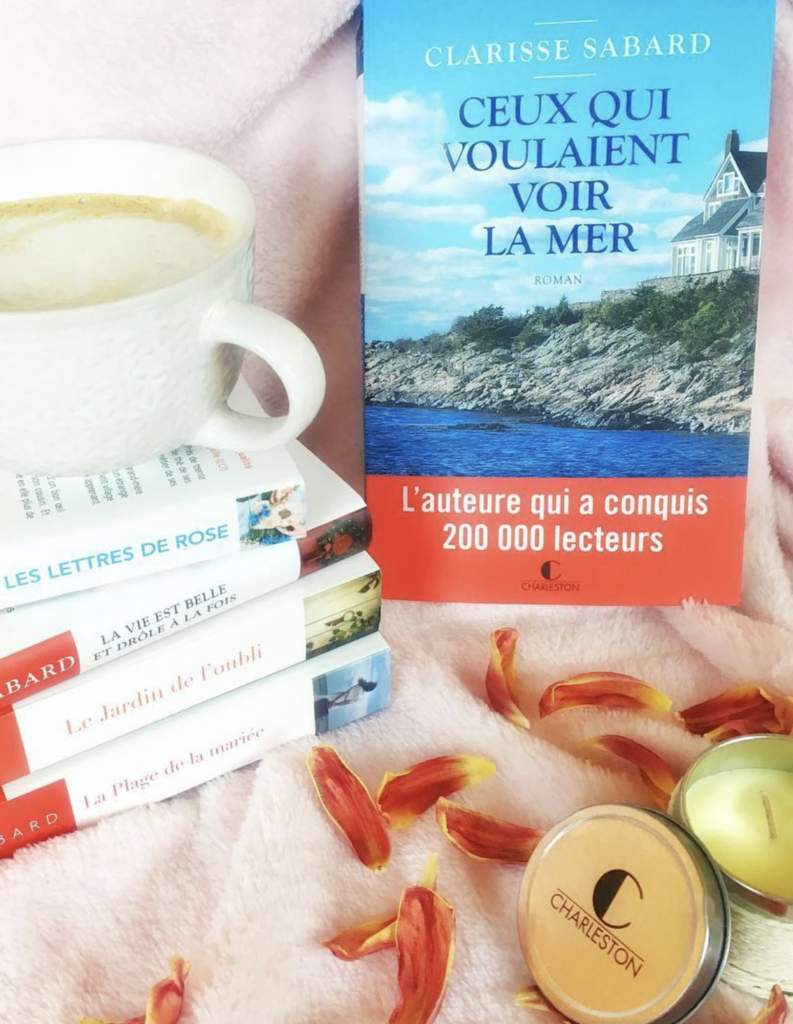 Le roman Ceux qui voulaient voir la mer de Clarisse Sabard est photographié à côté d'une tasse de café, de plusieurs romans de l'autrice, de bougies Charleston et de pétales de fleurs.