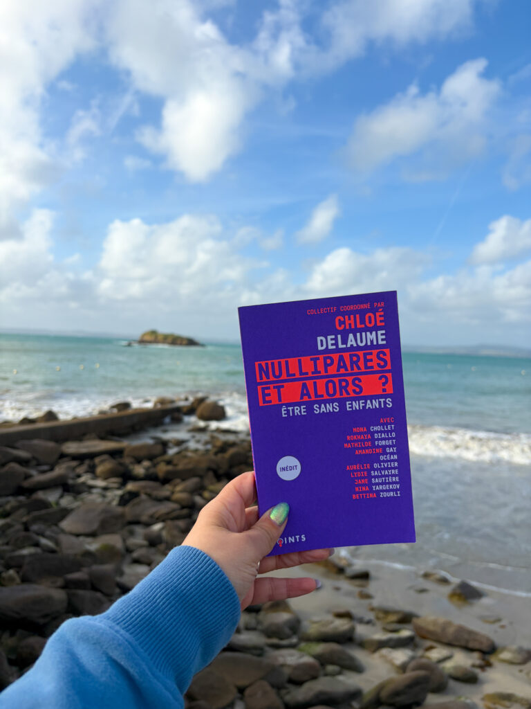 Le livre Nullipares et alors ? Être sans enfants, coordonné par Chloé Delaume, tenu à la main devant la plage de Tréboul à Douarnenez, en Bretagne.