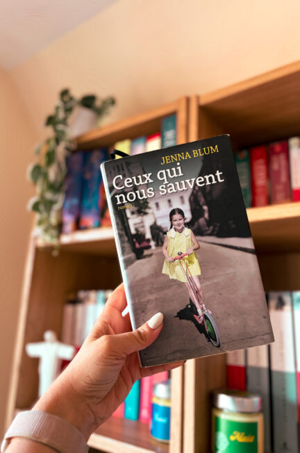 Une main tient le roman Ceux qui nous sauvent de Jenna Blum devant une bibliothèque en bois aux tranches de livres colorés avec une plante verte tombante posée sur l'étagère du dessus.