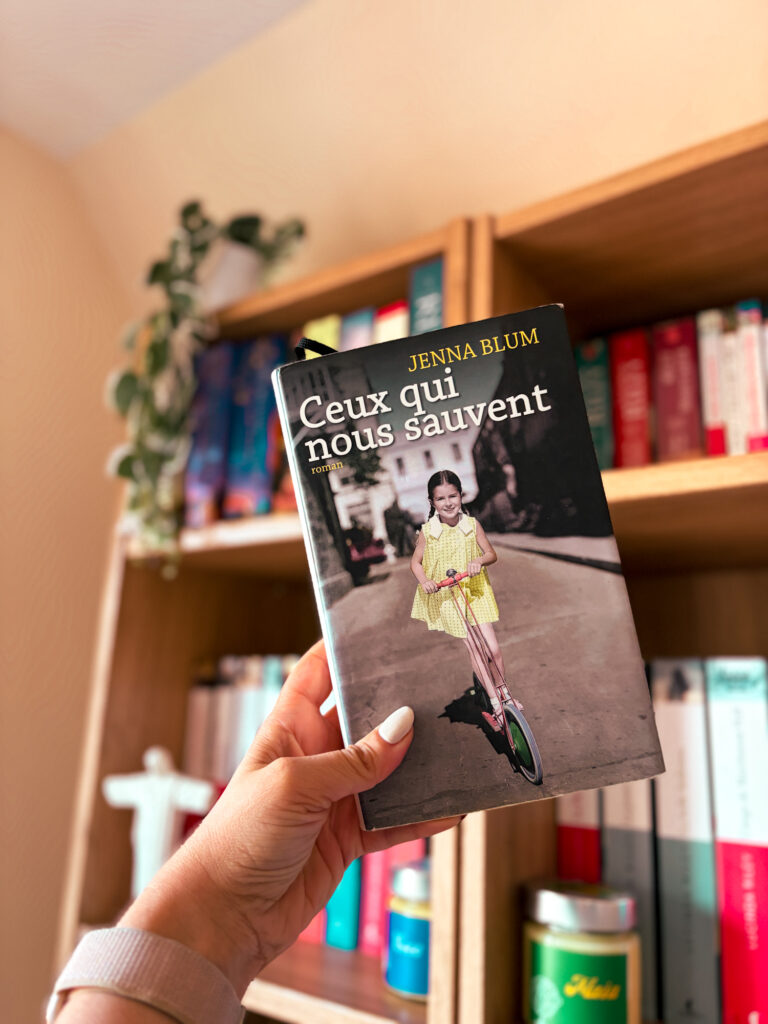 Une main tient le roman Ceux qui nous sauvent de Jenna Blum devant une bibliothèque en bois aux tranches de livres colorés avec une plante verte tombante posée sur l'étagère du dessus.