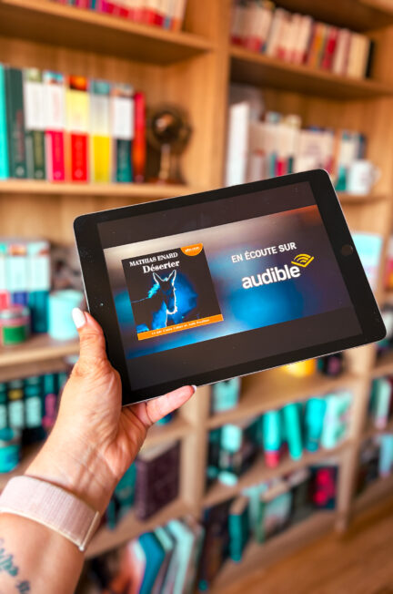 Le livre audio Déserter de Mathias Enard affiché sur un iPad tenu à la main devant une bibliothèque en bois aux tranches de livres colorées.