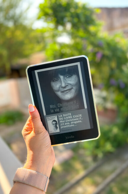 Une liseuse Kindle affichant la couverture de Moi Christiane F. la vie malgré tout de Christiane Felscherinow, tenue à la main devant un jardin baigné par la lumière du soleil.