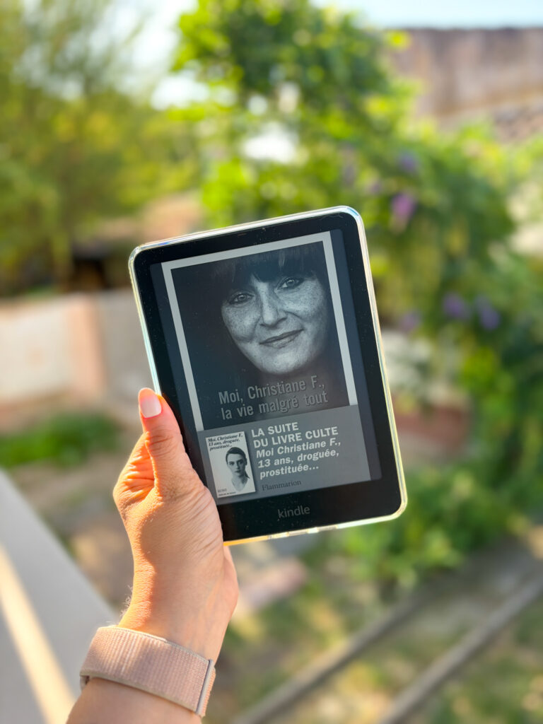 Une liseuse Kindle affichant la couverture de Moi Christiane F. la vie malgré tout de Christiane Felscherinow, tenue à la main devant un jardin baigné par la lumière du soleil.