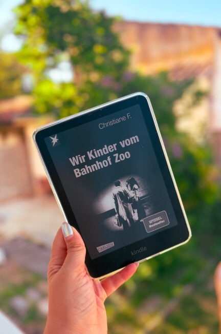 Une liseuse Kindle affichant la couverture de Wir Kinder vom Bahnhof Zoo de Christiane F., la version originale allemande de Moi Christiane F. 13 ans droguée prostituée, tenue à la main devant un jardin au coucher du soleil.