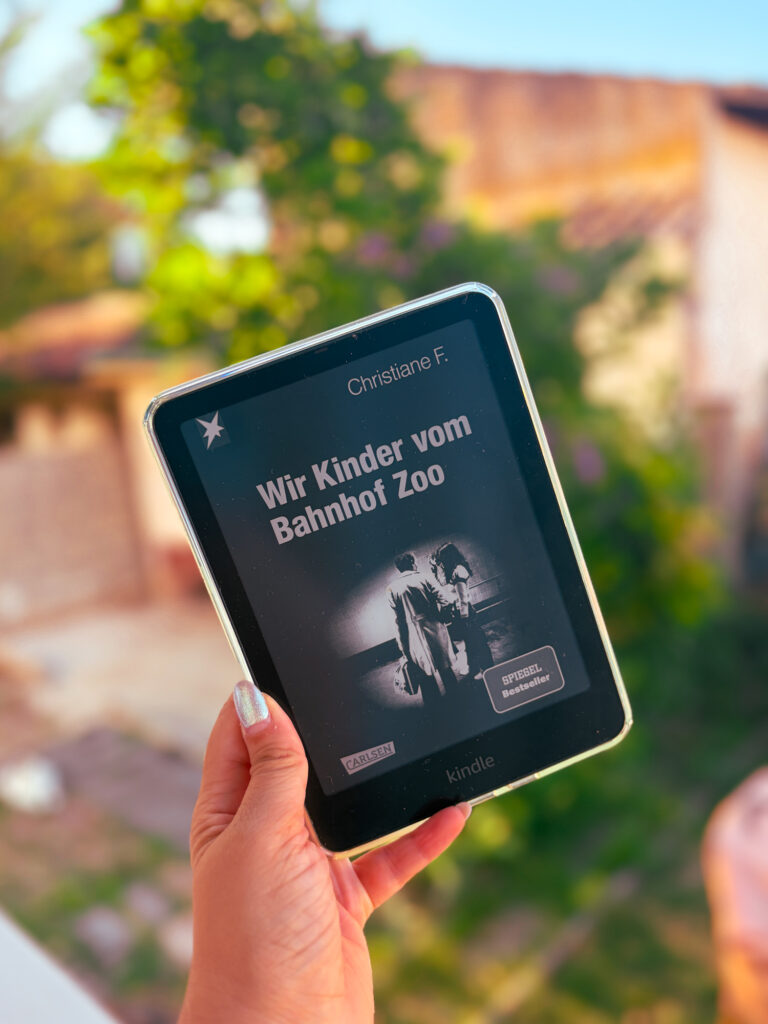 Une liseuse Kindle affichant la couverture de Wir Kinder vom Bahnhof Zoo de Christiane F., la version originale allemande de Moi Christiane F. 13 ans droguée prostituée, tenue à la main devant un jardin au coucher du soleil.