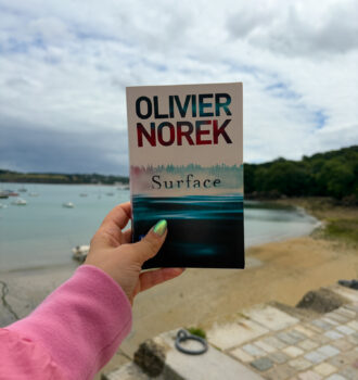 Le roman Surface d’Olivier Norek tenu à la main devant le port de Douarnenez, en Bretagne, avec la baie et les bateaux en arrière-plan.