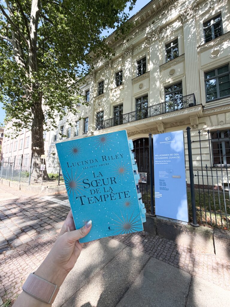 Main qui tient le tome 2 des sept soeurs de Lucinda Riley devant la porte cochère de la maison Schumann de Leipzig