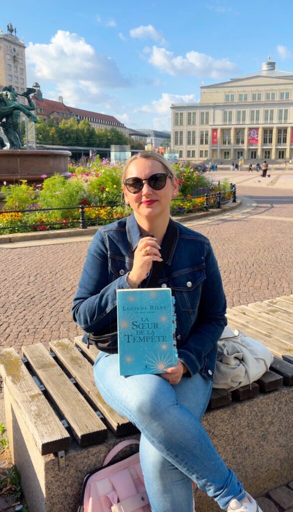 Sorbet-Kiwi assise sur un banc en pierre, tenant le tome 2 des sept soeurs de Lucinda Riley, avec en arrière plan l'opéra du Augustusplatz de Leipzig
