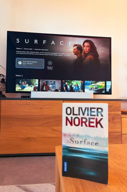 La série Surface affichée sur France TV sur une TV en arrière-plan, le roman d'Olivier Norek est posé sur une table en bois en premier plan