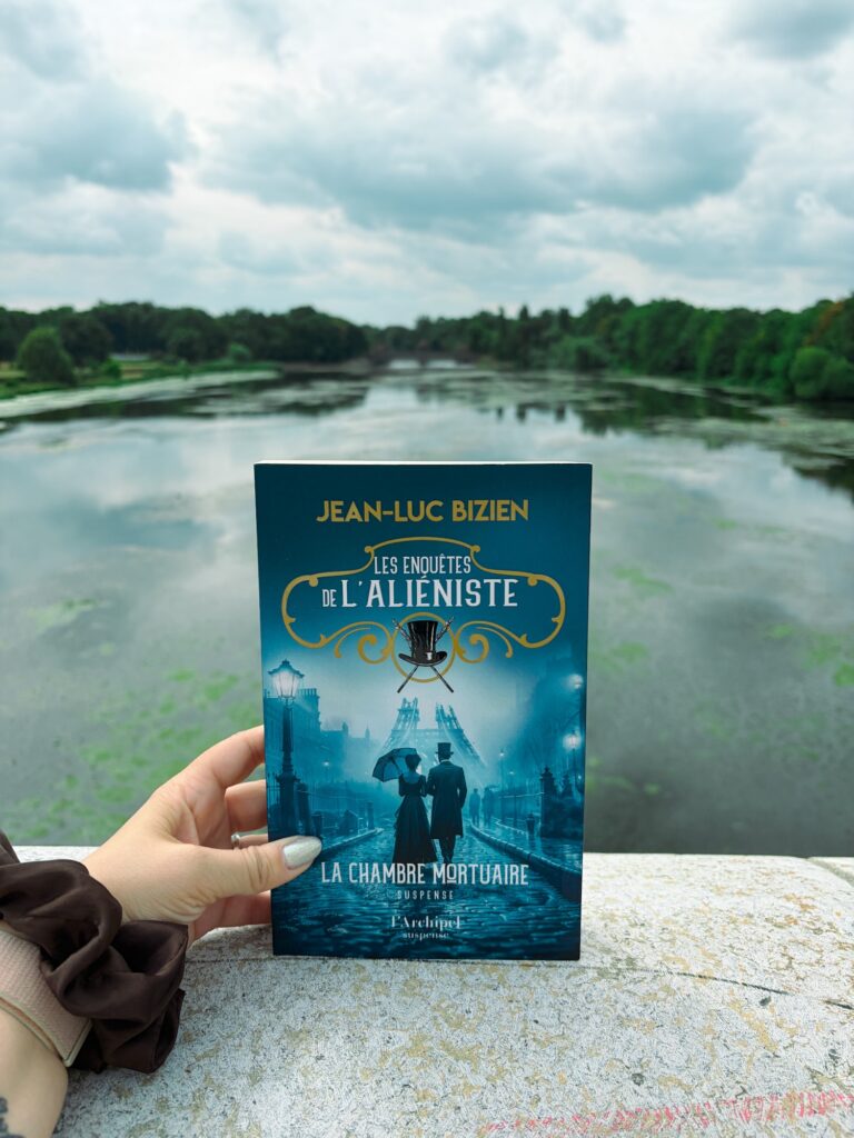 Livre La chambre mortuaire de Jean-Luc Bizien tenu à la main devant un plan d’eau bordé d’arbres à Leipzig.