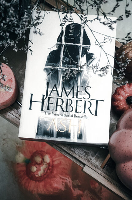 Photographie du roman Ash de James Herbert, dans son édition originale en anglais. Le livre est posé sur une table en bois, entouré de petites citrouilles décoratives orangées et rosées ainsi que de branches fleuries. Sa couverture blanche présente une silhouette sombre derrière une fenêtre aux carreaux irréguliers. Le titre Ash et le nom de James Herbert apparaissent en grandes lettres. Les tons désaturés et les ombres renforcent l’atmosphère gothique de cette mise en scène automnale.