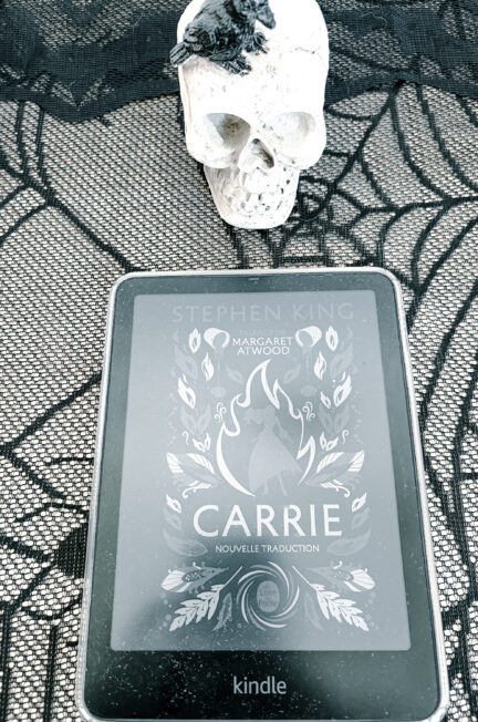 Photographie du roman Carrie de Stephen King sur une liseuse Kindle, posée sur une nappe en dentelle noire, sous un crâne décoratif blanc.