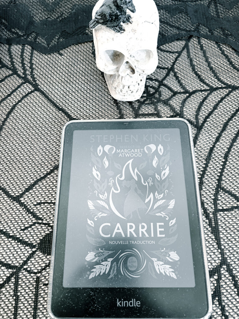 Photographie du roman Carrie de Stephen King sur une liseuse Kindle, posée sur une nappe en dentelle noire, sous un crâne décoratif blanc.