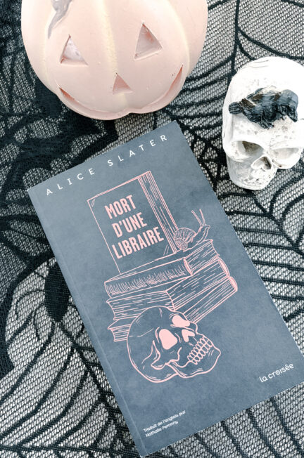 Photographie du roman Mort d'une libraire d'Alice Slater, publié aux éditions La Croisée. Le livre repose sur une dentelle noire à motifs de toiles d'araignée. Il est accompagné d'une citrouille décorative rose pâle et d'un crâne blanc sur lequel se trouve une petite souris noire. La composition évoque l'univers d'Halloween et accompagne une chronique littéraire consacrée à ce thriller psychologique.