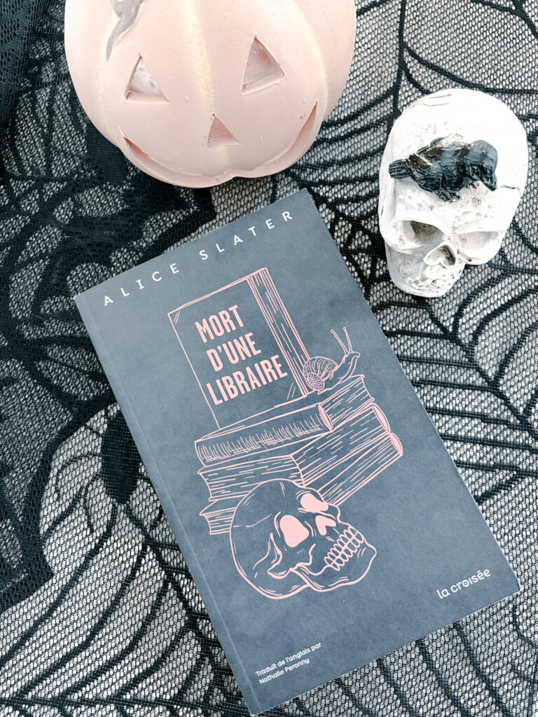 Photographie du roman Mort d'une libraire d'Alice Slater, publié aux éditions La Croisée. Le livre repose sur une dentelle noire à motifs de toiles d'araignée. Il est accompagné d'une citrouille décorative rose pâle et d'un crâne blanc sur lequel se trouve une petite souris noire. La composition évoque l'univers d'Halloween et accompagne une chronique littéraire consacrée à ce thriller psychologique.