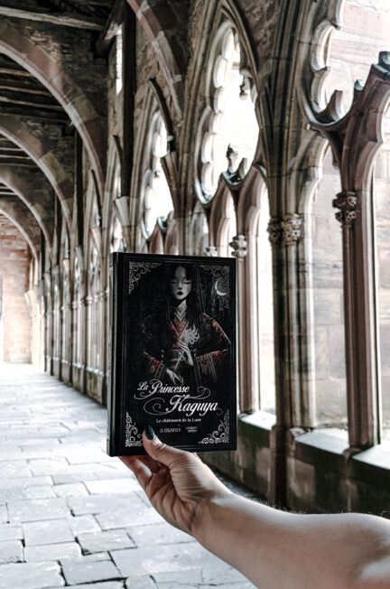 Photographie du roman La Princesse Kaguya : Le châtiment de la Lune de Dimitri Olivier, publié aux éditions Punchlines. Le livre est tenu à bout de bras devant les arcades gothiques du cloître de l’abbatiale Saints-Pierre-et-Paul à Wissembourg, en Alsace. La couverture noire représente une femme vêtue d’un kimono sombre aux motifs rouges, entourée de décorations florales. Les colonnes sculptées, les fenêtres gothiques et la perspective du cloître constituent l’arrière-plan de cette photographie.