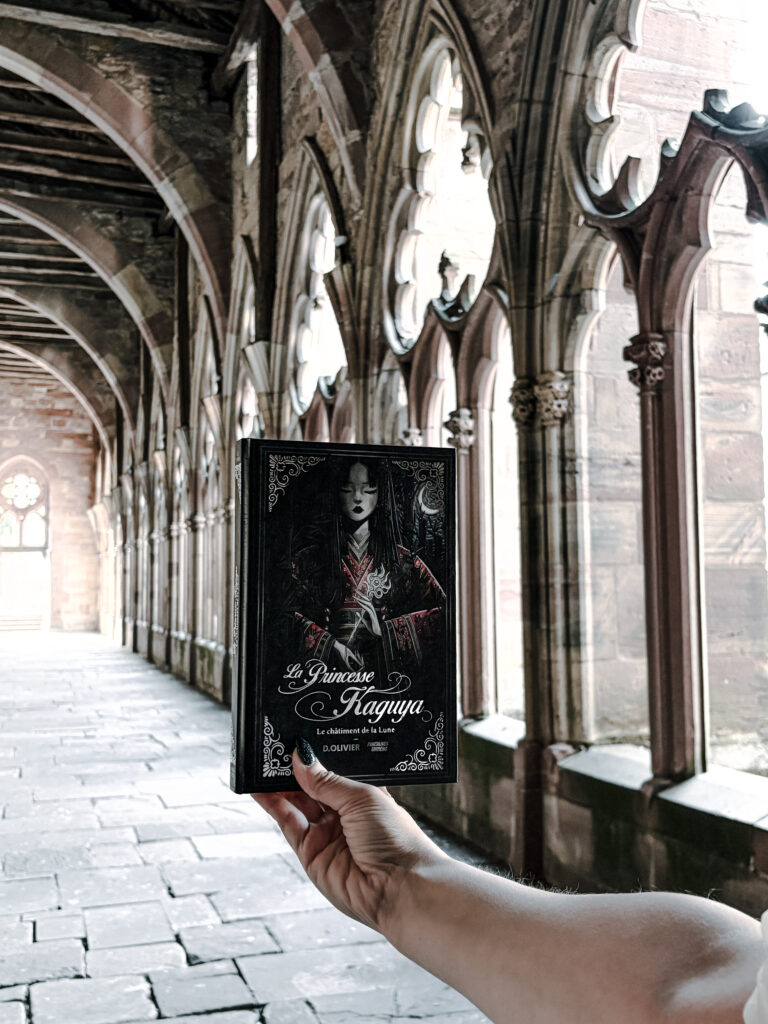 Photographie du roman La Princesse Kaguya : Le châtiment de la Lune de Dimitri Olivier, publié aux éditions Punchlines. Le livre est tenu à bout de bras devant les arcades gothiques du cloître de l’abbatiale Saints-Pierre-et-Paul à Wissembourg, en Alsace. La couverture noire représente une femme vêtue d’un kimono sombre aux motifs rouges, entourée de décorations florales. Les colonnes sculptées, les fenêtres gothiques et la perspective du cloître constituent l’arrière-plan de cette photographie.