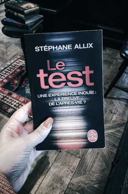 Photographie du livre Le test de Stéphane Allix, publié aux éditions Le Livre de Poche. Une main aux ongles orange tient le roman au premier plan. Sa couverture sombre présente le titre en grandes lettres rouges, accompagné du sous-titre « Une expérience inouïe : la preuve de l’après-vie ? ». En arrière-plan, on distingue un parquet en bois, un tapis à motifs géométriques, une petite pile de livres et du mobilier sombre. Cette photographie prise à europa-Park en 2021 accompagne une chronique littéraire consacrée à l’ouvrage de Stéphane Allix.