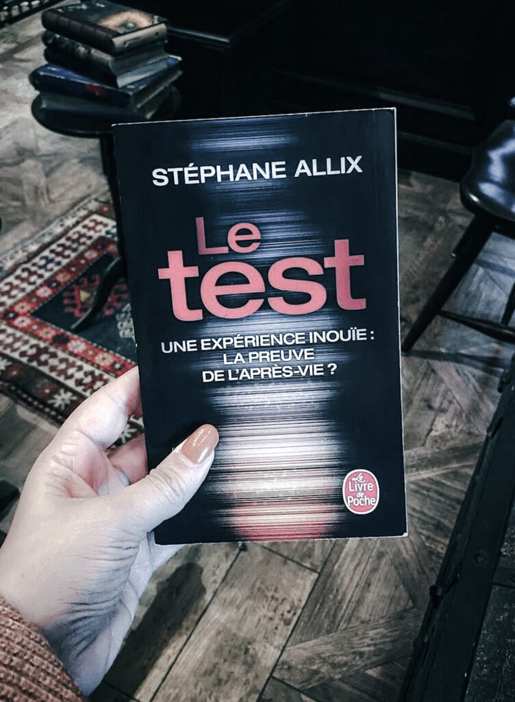 Photographie du livre Le test de Stéphane Allix, publié aux éditions Le Livre de Poche. Une main aux ongles orange tient le roman au premier plan. Sa couverture sombre présente le titre en grandes lettres rouges, accompagné du sous-titre « Une expérience inouïe : la preuve de l’après-vie ? ». En arrière-plan, on distingue un parquet en bois, un tapis à motifs géométriques, une petite pile de livres et du mobilier sombre. Cette photographie prise à europa-Park en 2021 accompagne une chronique littéraire consacrée à l’ouvrage de Stéphane Allix.