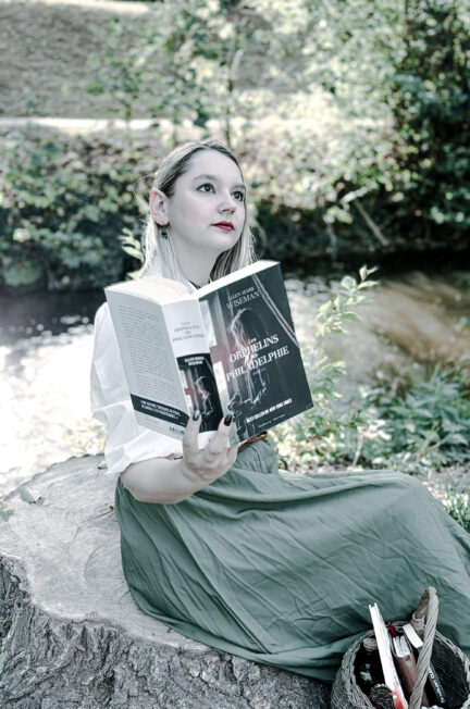 Photographie littéraire en extérieur représentant une lectrice blonde vêtue d’un chemisier blanc et d’une longue jupe verte, assise sur une souche d’arbre près d’un cours d’eau. Elle tient ouvert le roman Les Orphelins de Philadelphie d’Ellen Marie Wiseman, publié aux éditions Faubourg Marigny. Un panier contenant des livres est posé à ses côtés. La végétation et la lumière naturelle composent un décor paisible.