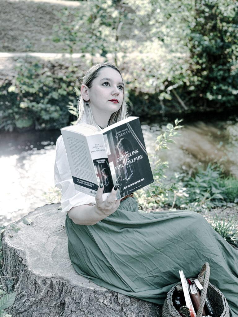 Photographie littéraire en extérieur représentant une lectrice blonde vêtue d’un chemisier blanc et d’une longue jupe verte, assise sur une souche d’arbre près d’un cours d’eau. Elle tient ouvert le roman Les Orphelins de Philadelphie d’Ellen Marie Wiseman, publié aux éditions Faubourg Marigny. Un panier contenant des livres est posé à ses côtés. La végétation et la lumière naturelle composent un décor paisible.
