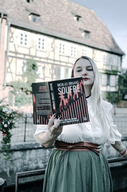 Photographie d’une femme blonde aux cheveux longs, vêtue d’une blouse blanche et d’une jupe verte, tenant ouvert le thriller Soufre de Nicolas Druart, publié aux éditions HarperCollins Noir. Elle se tient devant une grande maison alsacienne à colombages, près d’une rambarde métallique. La couverture noire et rouge du roman est bien visible au premier plan. Cette photographie accompagne une chronique littéraire consacrée au roman de Nicolas Druart.