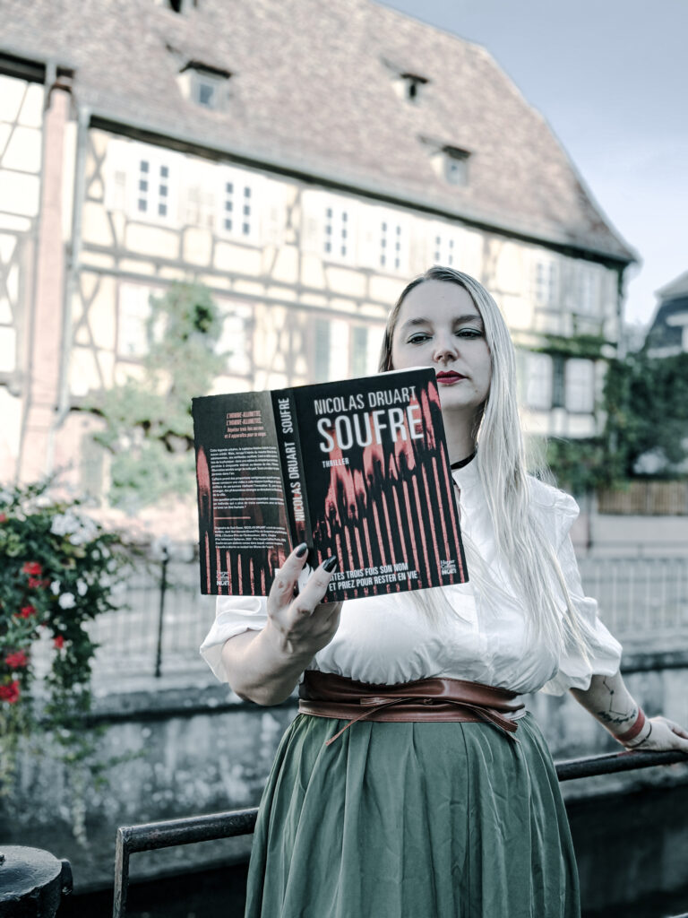 Photographie d’une femme blonde aux cheveux longs, vêtue d’une blouse blanche et d’une jupe verte, tenant ouvert le thriller Soufre de Nicolas Druart, publié aux éditions HarperCollins Noir. Elle se tient devant une grande maison alsacienne à colombages, près d’une rambarde métallique. La couverture noire et rouge du roman est bien visible au premier plan. Cette photographie accompagne une chronique littéraire consacrée au roman de Nicolas Druart.